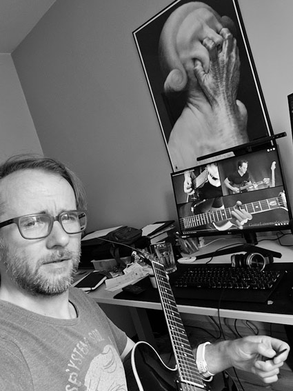 Maciej Lesiak - A black and white selfie in a home office setting. The subject is wearing glasses and has a neatly trimmed beard, dressed in a casual t-shirt with a graphic print. In the frame, there's an acoustic guitar visible in the foreground. Behind him, there's a computer setup with dual monitors - one showing what appears to be a guitar tutorial video, and above it, an artistic black and white photograph of what appears to be a human form. The workspace suggests a music practice/learning environment combined with a home office setup.